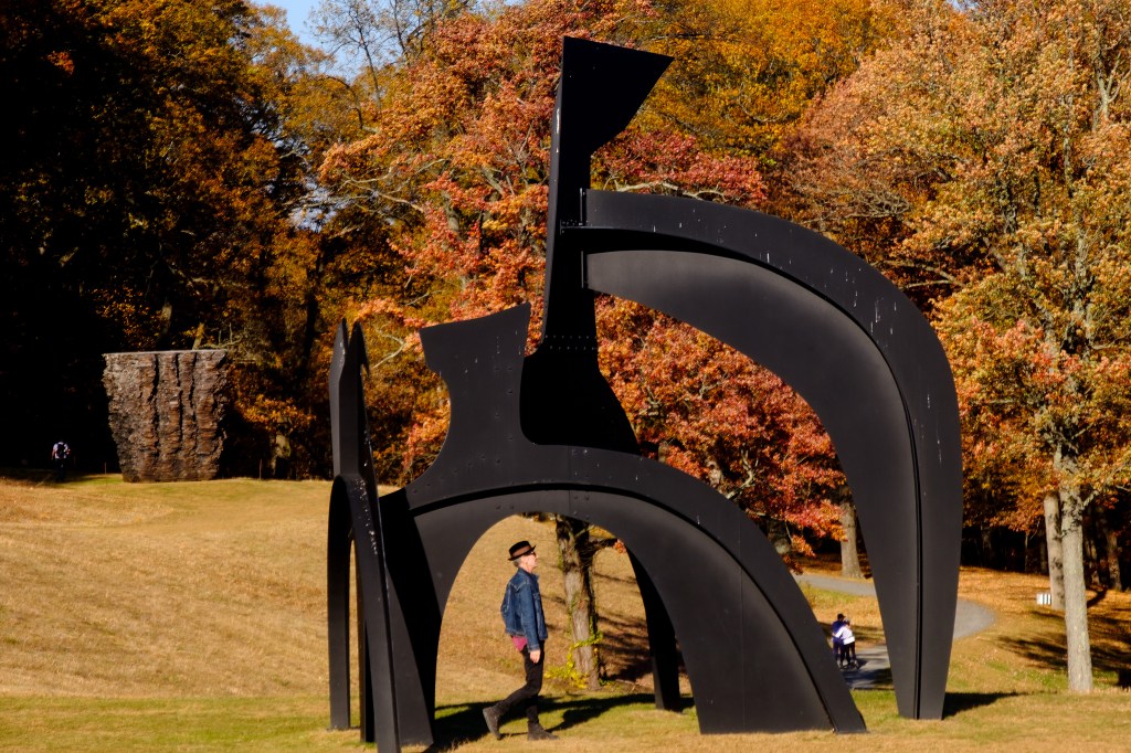 A man walking under Alexander Calder's Black Flag sculpture at the Storm King Art Center amid autumn leaves